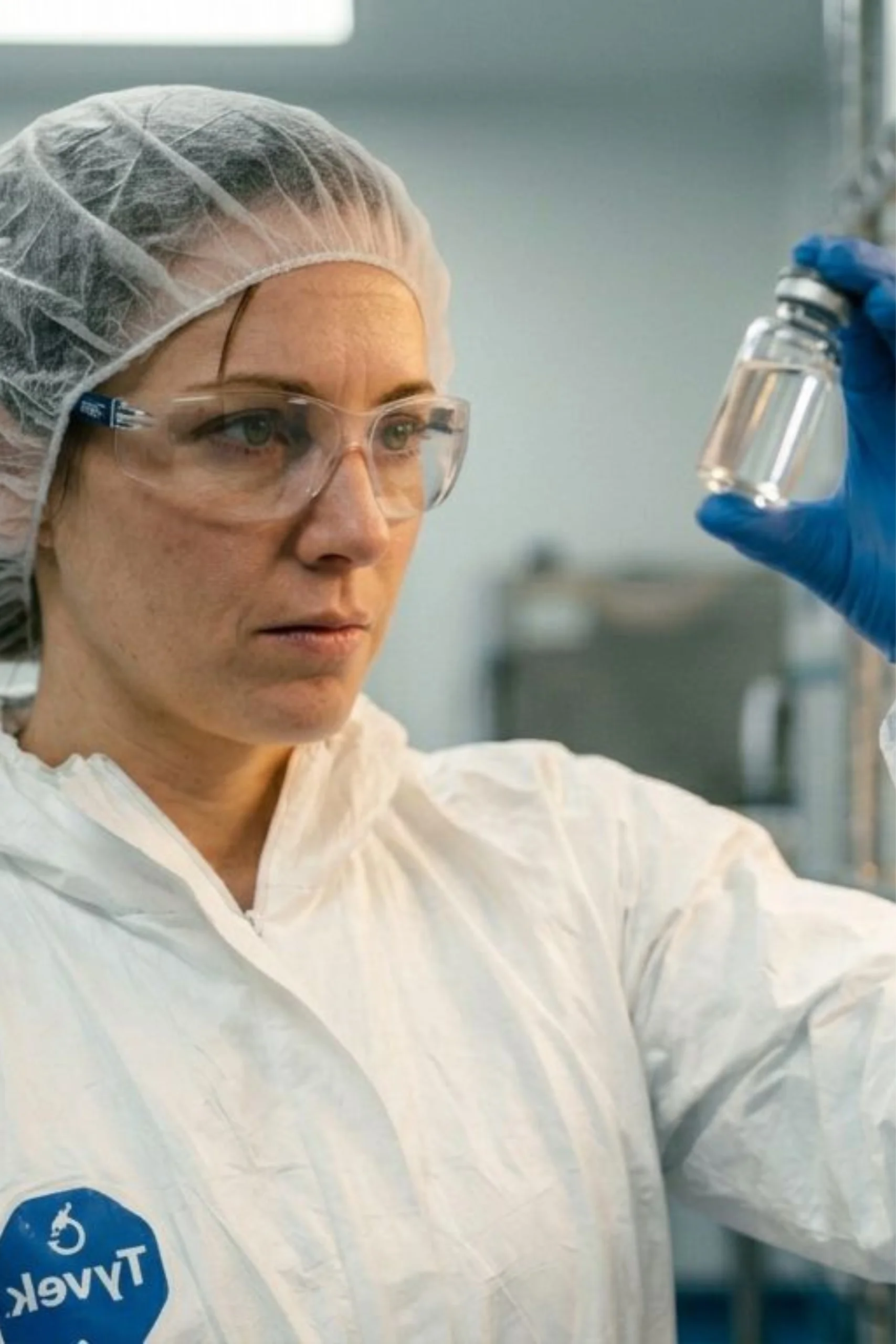 HerbiLabs female quality control technician in Tyvek suit and hairnet holding clear glass vial up to light for inspection in pharmaceutical cleanroom