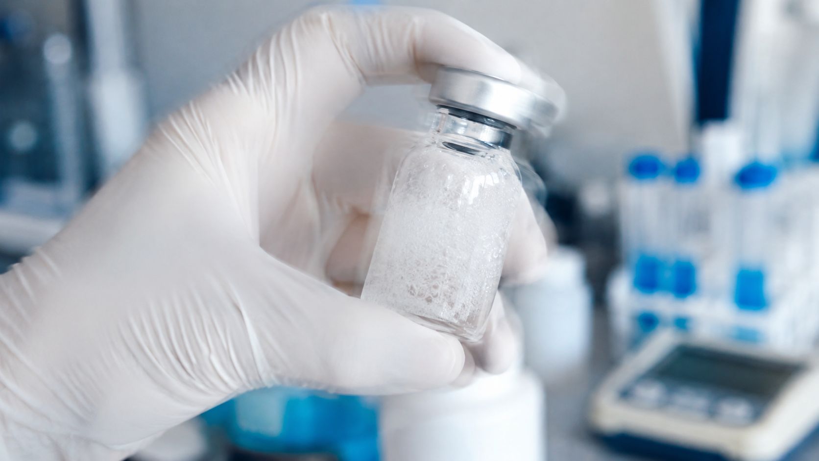 A scientist in white gloves holding a glass vial containing a white substance for mixing peptides.
